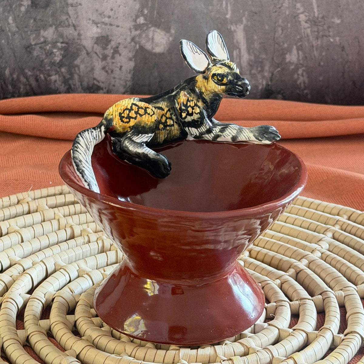 rooibos red ceramic mini raised bowl with a hand painted african wild dog crouched on the rim, ears wide and alert